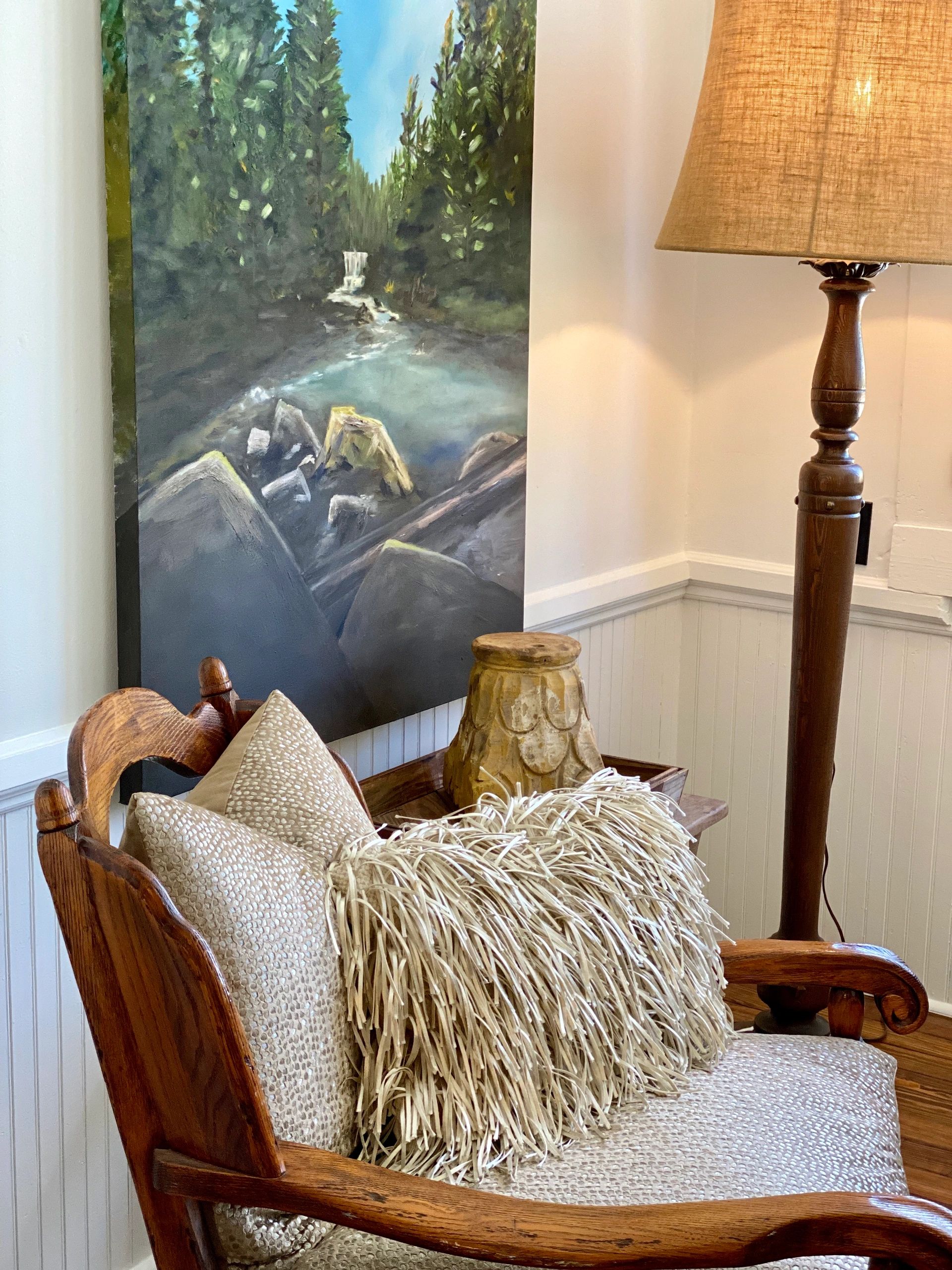 Cozy corner with wooden chair, textured pillows, standing lamp, and forest landscape painting